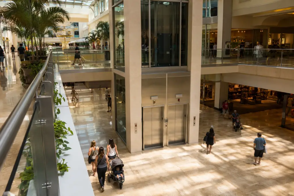Multi-level Florida retail shopping center elevator bank with stainless steel doors and high-traffic shopper activity, illustrating retail elevator maintenance standards compliance needs.