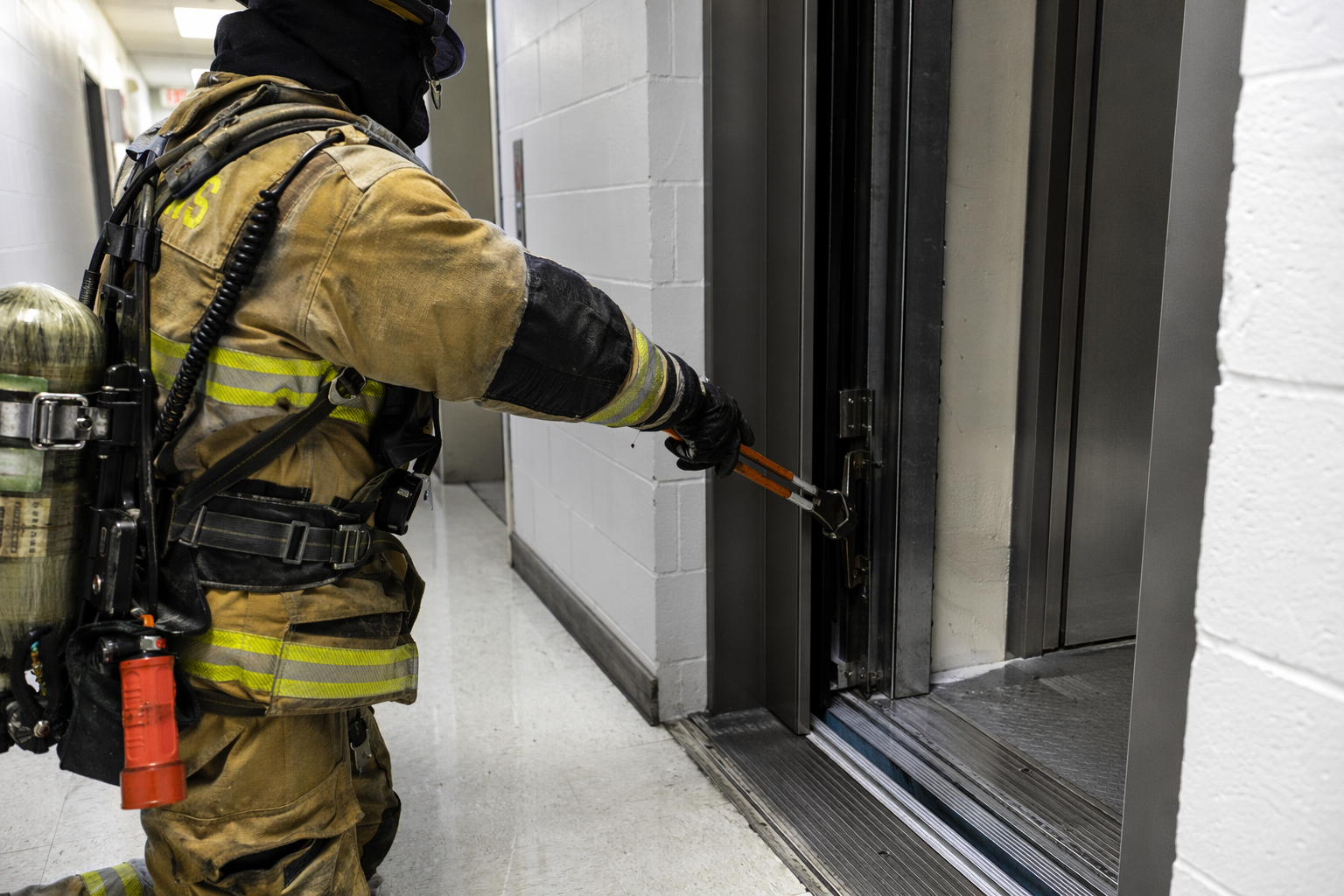 Firefighter using a manual door release tool to open a stuck elevator door during an entrapment rescue in a Florida commercial building hallway.