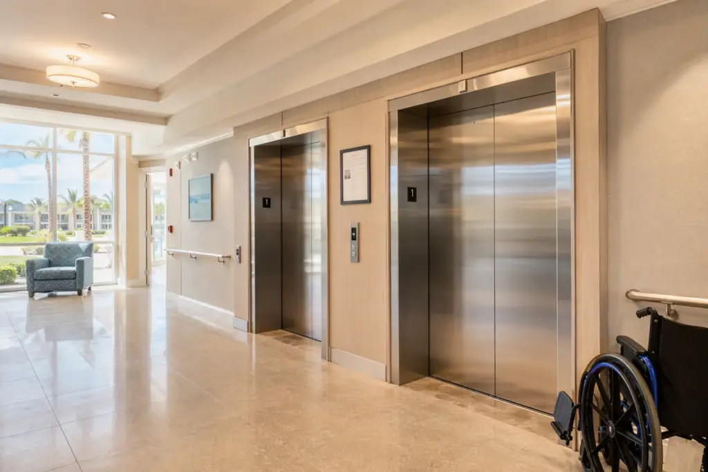 ADA-compliant elevator lobby in a Florida assisted living facility with wide stainless-steel doors, braille call buttons, and Certificate of Operation posted on the wall