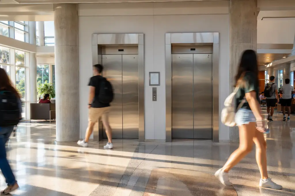 University lobby in Florida showing stainless-steel elevator doors with a DBPR certificate of operation mounted on the wall beside the call panel, natural afternoon light