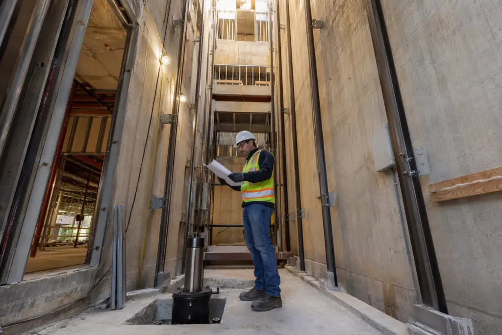 Licensed elevator mechanic reviewing permit drawings inside a commercial hoistway under construction in a Florida office building during new elevator installation