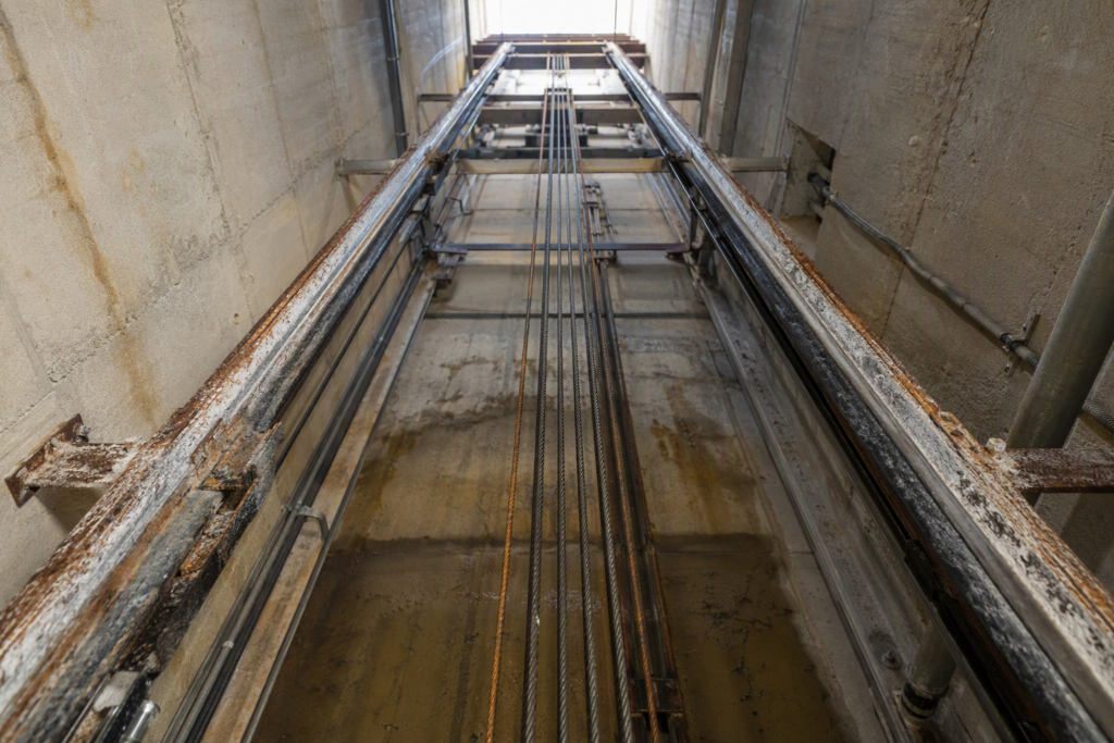 Open elevator hoistway in a Florida coastal building showing corroded guide rails and salt deposits on steel components caused by humidity and salt air exposure